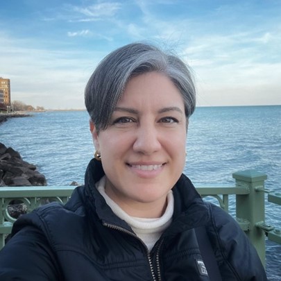 Smiling woman by the water with a railing and shoreline behind her.